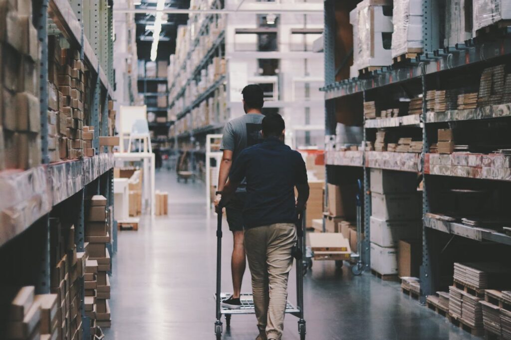 pexels-photo-1797428 Two men maneuver a trolley in a large warehouse filled with boxes and shelves.
